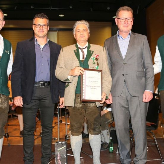 Fünf Männer in formeller Kleidung stehen auf der Bühne und halten einen Preis. Ein Mann in der Mitte hält ein gerahmtes Zertifikat, während andere traditionelle deutsche Kleidung tragen.