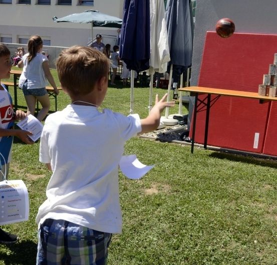Kinder nehmen an einem Spiel teil, bei dem ein Ball und ein Stapel von Dosen an einer roten Wand beteiligt sind. Ein Junge wirft den Ball.