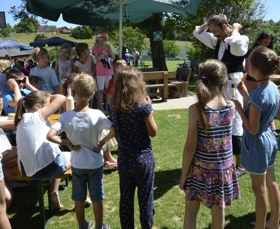 Eine Gruppe von Kindern und Erwachsenen versammelt sich im Freien. Einige sitzen an Tischen, während andere stehen und einem Mann zuhören. Der Bereich ist von Schirmen beschattet und von Bäumen und einem Rasenfeld umgeben.