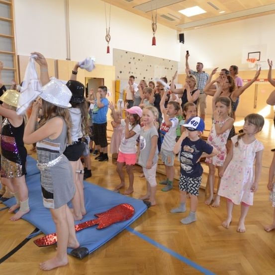 Eine Gruppe von Kindern und Erwachsenen nimmt in einer Turnhalle an einer Tanzaufführung teil, einige tragen Hüte und Turnschuhe. Ein Erwachsener hält ein Handy. An der Wand ist ein Basketballkorb zu sehen.