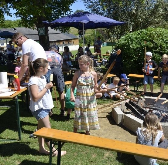 Eine Gruppe von Kindern und Erwachsenen versammelt sich um ein Feuerloch, wobei einige Lebensmittel kochen und andere mit Stöcken spielen. In der Nähe sind Tische und Stühle zum Essen aufgestellt, und ein blauer Regenschirm spendet Schatten. Die Umgebung scheint ein Park mit Bäumen und einem Gebäude in der Ferne zu sein.