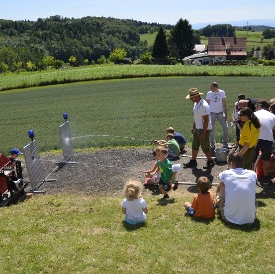 Eine Gruppe von Menschen, einschließlich Kindern, versammelt sich um einen roten Traktor auf einer grasigen Wiese, wobei ein Mann einen Wasserschlauch benutzt. Dahinter liegt eine weitläufige grüne Wiese mit Bäumen und Häusern.