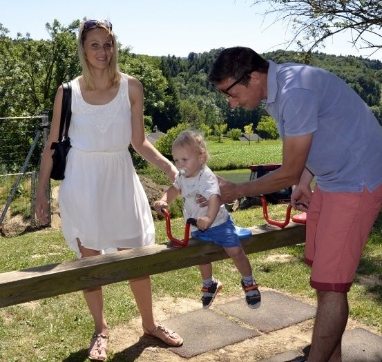 Eine Frau, ein Mann und ein Kleinkind sind in einem Park und spielen auf einer Wippe. Das Kleinkind saugt an einem Schnuller.