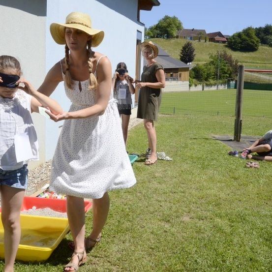 Eine Gruppe von Kindern spielt an einem sonnigen Tag draußen, wobei ein Mädchen die Augen verbunden hat. Eine Frau in einem Hut führt sie. In der Nähe sitzt ein Junge auf dem Gras. Dahinter steht ein Gebäude mit einem Zaun und einem Spielplatz.