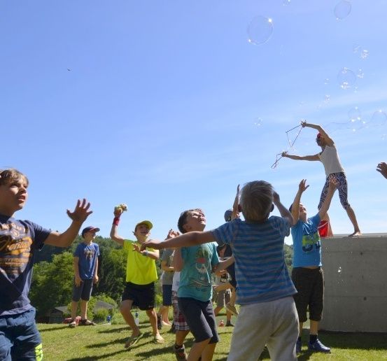 Eine Gruppe von Kindern spielt mit Seifenblasen an einem sonnigen Tag in einem grünen Außenbereich mit einer Wand und Bäumen im Hintergrund.