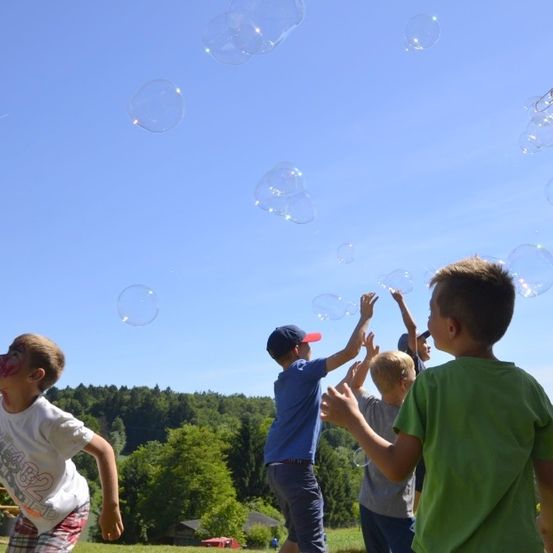 Kinder spielen im Freien in einem Park und pusten Seifenblasen in einen klaren blauen Himmel. Bäume und ein Gebäude in der Ferne sind sichtbar.