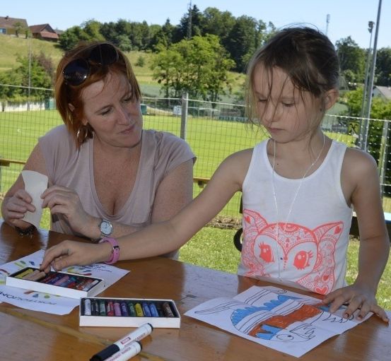 Eine Frau und ein junges Mädchen sitzen draußen an einem Tisch und machen Kunst. Das Mädchen hat Buntstifte und ein Malbuch. Sie sind in der Nähe eines Rasens mit einem Zaun.