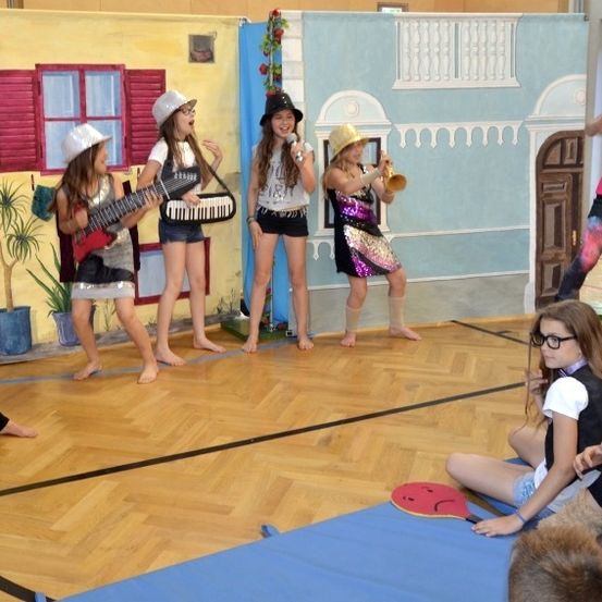 Eine Gruppe von Kindern führt eine musikalische Darbietung auf einer Bühne auf. Vier Mädchen spielen Musikinstrumente und ein weiteres Mädchen singt. Ein Topf mit einer Pflanze und ein bemalter Hintergrund sind im Hintergrund zu sehen.