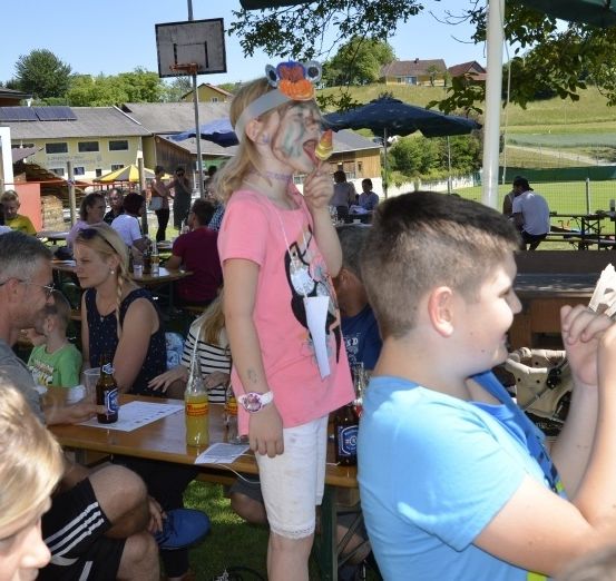 Eine Gruppe von Kindern und Erwachsenen versammelt sich bei einer Veranstaltung im Freien. Ein Mädchen isst einen Lutscher und ein Junge in der Nähe hält ein Papier. Tische mit Getränken und Essen sind aufgestellt. Die Veranstaltung befindet sich in der Nähe eines Basketballplatzes, mit Gebäuden und Grünflächen im Hintergrund.