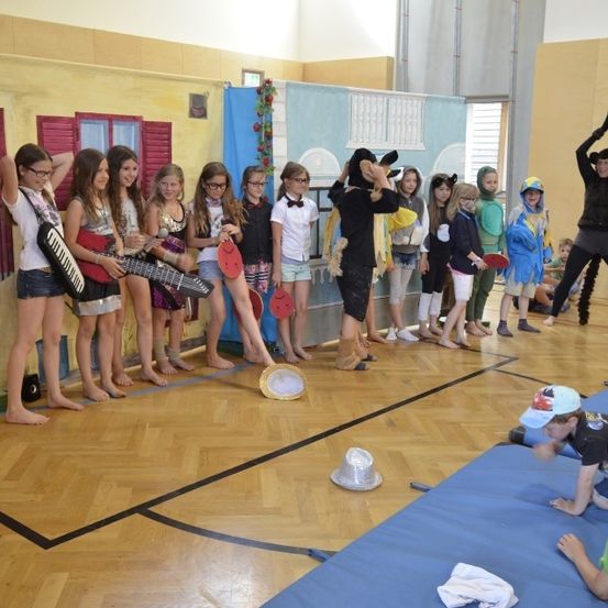 Eine Gruppe von Kindern in verschiedenen Kostümen steht in einer Turnhalle, eines hält eine Gitarre und andere haben Requisiten, mit einer Person, die Yoga in der Hintergrund macht.