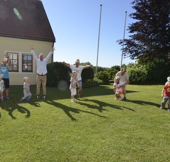 Eine Gruppe von Menschen steht auf einem Rasen, einige mit Hüten, und feiert. Dahinter ein Haus und Bäume.