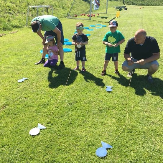 Vier Personen, darunter zwei Kinder, sind auf einem Rasenfeld bei einer Outdoor-Aktivität. Sie stehen und knien und halten Schnüre mit fischförmigen Objekten. Dahinter befinden sich mehrere blaue Scheiben und eine Metallkonstruktion.