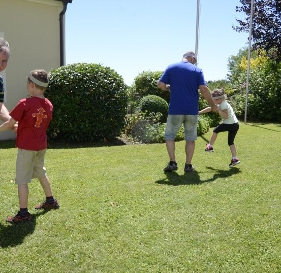 Eine Gruppe von Menschen, einschließlich Erwachsener und Kindern, halten sich an den Händen in einem Rasenbereich, mit einem Haus und Hecken im Hintergrund.