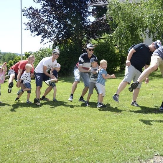 Eine Gruppe von Erwachsenen und Kindern steht auf einem Rasen unter einem klaren Himmel und nimmt an einem Staffellauf teil. Ein Erwachsener lehnt sich vor, während Kinder neben ihm laufen.