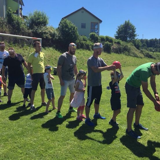Eine Gruppe von Erwachsenen und Kindern mit verbundenen Augen und roten Bällen auf einem Rasenfeld mit einem Fußballtor im Hintergrund.