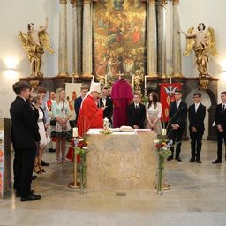 Eine religiöse Zeremonie mit einem Priester und vielen Menschen in einer Kirche. Hinter dem Priester befindet sich ein großes Gemälde und ein Tisch mit Blumen und Büchern.