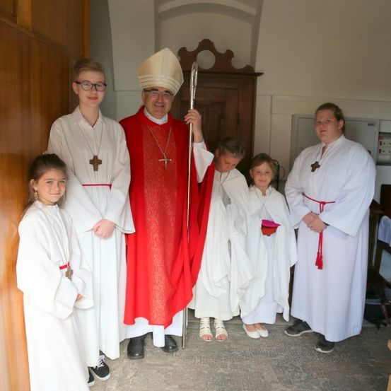 Eine Gruppe von Menschen, darunter ein Priester in einem roten Gewand, steht in einem Raum. Sie tragen alle weiße Gewänder, einige halten Bücher. Der Raum hat Holzschränke und eine weiße Wand.