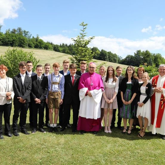 Eine Gruppe von Menschen steht auf einem Feld mit einem Priester und einem religiösen Amtsträger vor einem Haus und Bäumen.
