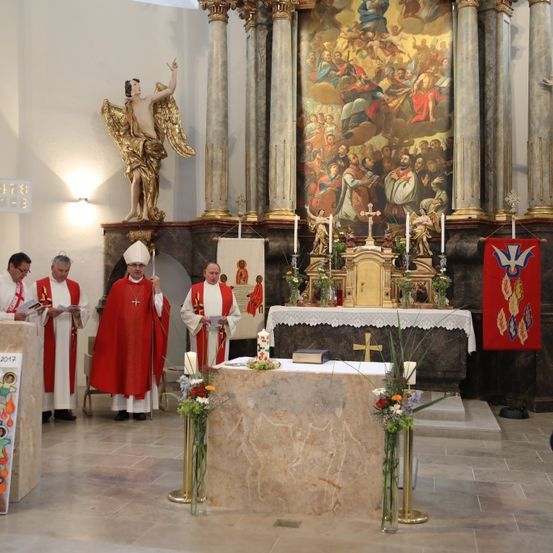 Mehrere Priester in roten Gewändern stehen am Altar in einer Kirche, möglicherweise bei einer Zeremonie. Hinter ihnen befindet sich ein großes Gemälde an der Wand, sowie mehrere Statuen und Banner.