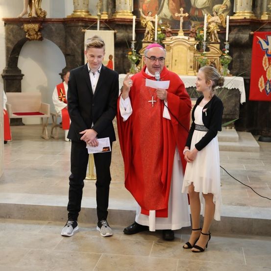 Ein Priester hält ein Mikrofon, während ein junger Junge und ein Mädchen neben ihm stehen, wahrscheinlich bei einer Kirchenveranstaltung.