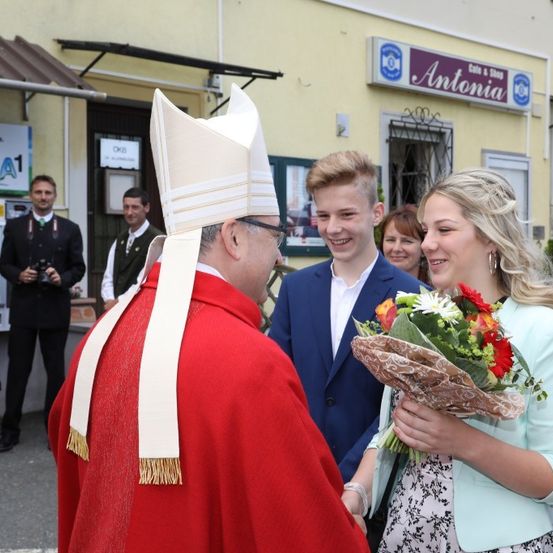Ein Priester in roten Gewändern begrüßt ein lächelndes Paar mit Blumen. Dahinter steht ein Mann mit einer Kamera in der Nähe eines Gebäudes mit dem Schild 'Antonia'.