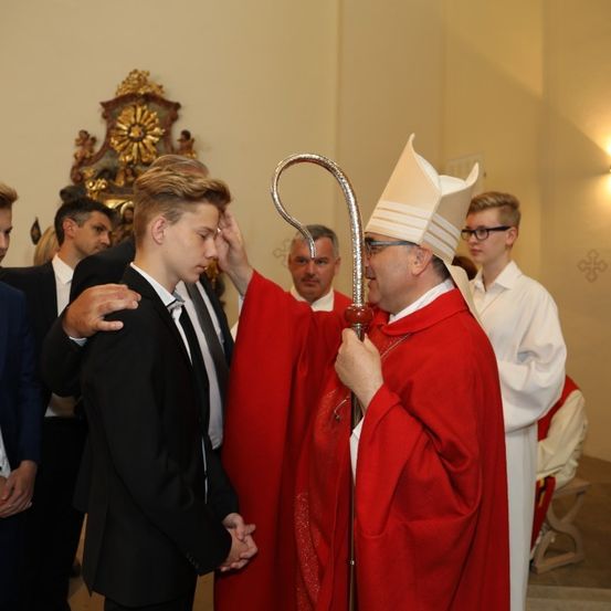 Ein Priester in roter zeremonieller Kleidung legt seine Hand auf den Kopf eines jungen Mannes in einer Kirchenumgebung. Ein weiterer Priester in Rot und mehrere andere Männer stehen hinter ihnen.