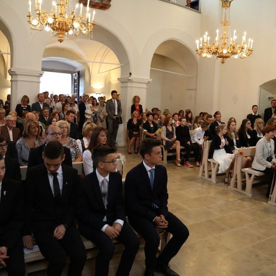 Eine große Gruppe von Menschen ist in einer Kirche versammelt, viele sitzen auf Bänken. Einige Personen stehen im Hintergrund. Zwei Kronleuchter hängen von der Decke.