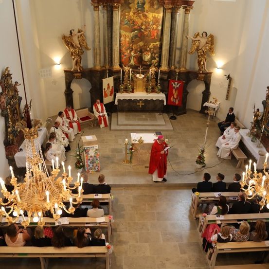 Ein Priester leitet einen Gottesdienst in einer Kirche mit Altar, Kerzen und Statuen. Die Leute sitzen auf Bänken und Kronleuchter hängen von der Decke.