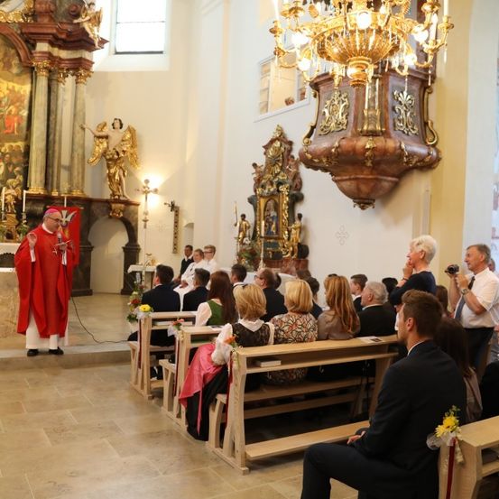 Ein Priester in einer roten Robe spricht zu einer Gemeinde in einer Kirche. Der Altar hat Gemälde und Kerzen. Menschen sitzen auf Bänken, und einige stehen.