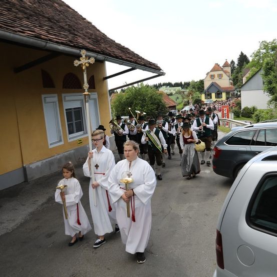 Eine Gruppe von Menschen in traditioneller Kleidung zieht in einer Prozession, Kreuze und Laternen tragend, mit einer Brassband hinter ihnen. Sie sind vor einem Gebäude, mit geparkten Autos in der Nähe.