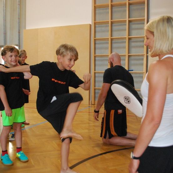 In einer Turnhalle lächelt eine Frau, während sie den Arm eines Jungen hält, und ein Junge übt einen Tritt. Ein anderer Junge schaut sie an. Ein Mann kniet und hält einen Pad. Ein hölzernes Bauwerk ist im Hintergrund.