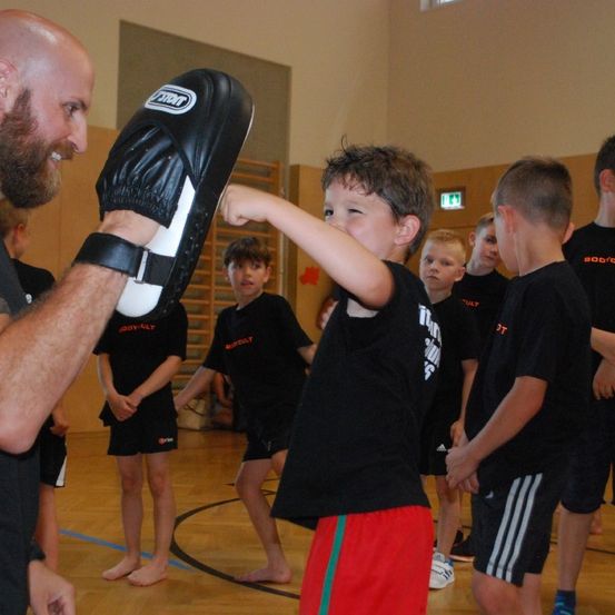 Ein Mann in einem Fitnessstudio hält eine schwarze Boxhandschuh-Polster für einen Jungen zum Schlagen. Der Junge zielt auf die Polster, während andere in schwarzen Uniformen zusehen.