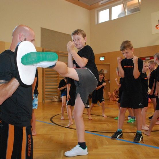 Ein junger Junge übt Kampfkunst, indem er eine von einem Mann gehaltene Polster tritt, in einer Turnhalle. Andere Kinder trainieren im Hintergrund.