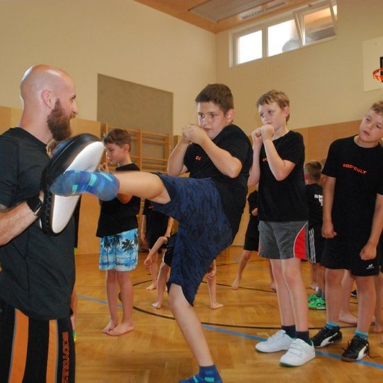 Ein Mann unterrichtet einen jungen Jungen, mit seinem Bein zu treten, in einer Turnhalle. Der Junge trägt Shorts und ein T-Shirt. Dahinter stehen andere Kinder und beobachten.