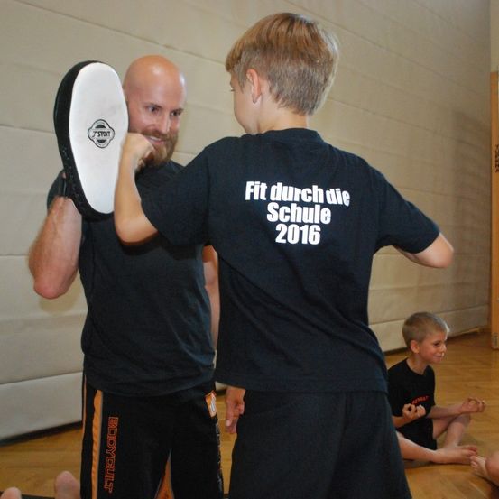 Ein Mann hält eine schwarz-weiße Polsterung, während ein Junge Kampfsport in einer Turnhalle übt. Der Junge trägt ein schwarzes T-Shirt mit dem Text 'Fit durch die Schule 2016'. Ein weiteres Kind sitzt hinter ihnen auf dem Boden.