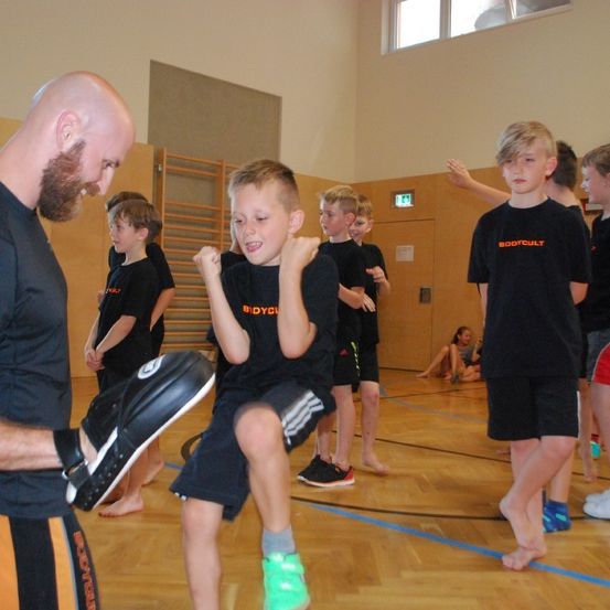 Eine Gruppe von Kindern in schwarzen T-Shirts und Shorts nimmt an einer körperlichen Aktivität teil, möglicherweise Kampfsport. Sie befinden sich in einer Turnhalle mit Holzboden. Ein Mann unterrichtet sie, und sie stehen in einer Reihe. Im Hintergrund sitzt ein Mädchen auf dem Boden.