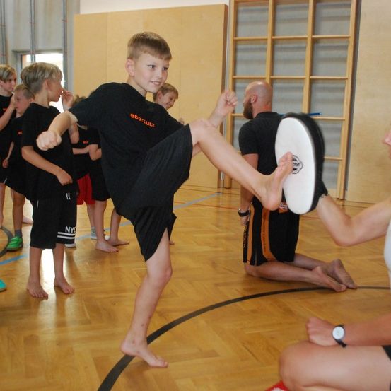 Ein Junge übt hohe Kicks in einer Turnhalle, während eine Frau ein Pad für ihn hält. Mehrere andere Jungen stehen in der Nähe und beobachten.
