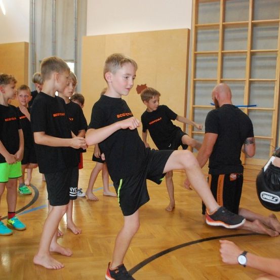 Eine Gruppe junger Jungen trainiert Kampfsport in einer Turnhalle. Sie tragen schwarze T-Shirts mit orangefarbenem Aufdruck. Ein Mann mit Tattoo und schwarzer Shorts unterrichtet sie.
