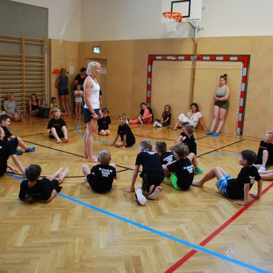 Ein Hallenbasketballfeld mit einer stehenden Frau, die wahrscheinlich eine Gruppe von Kindern in einem Kreis anweist, die identische schwarze T-Shirts tragen. Ein Basketballkorb ist an der Wand montiert, und einige Erwachsene sitzen und stehen in der Nähe.