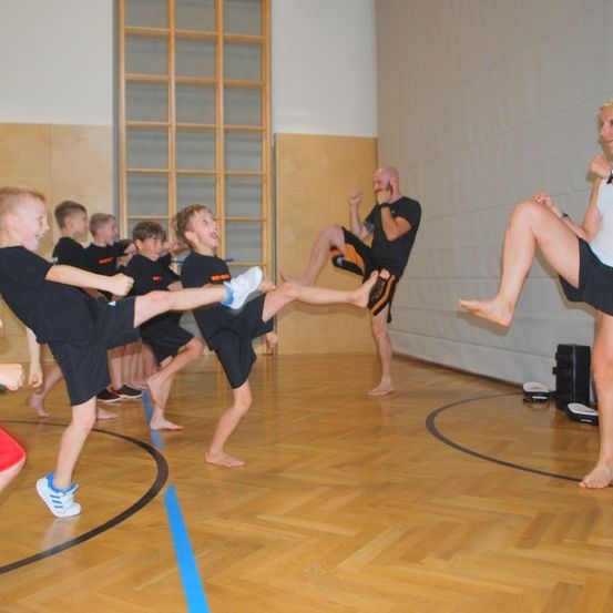 Mehrere Kinder in einer Turnhalle praktizieren Kampfkunst mit einem Ausbilder. Der Ausbilder demonstriert einen Kick.