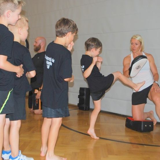 Eine Gruppe von Kindern und einer erwachsenen Frau übt Kampfsport in einem Fitnessstudio. Die Frau hält eine Polsterung, während ein Junge einen hohen Tritt ausführt.