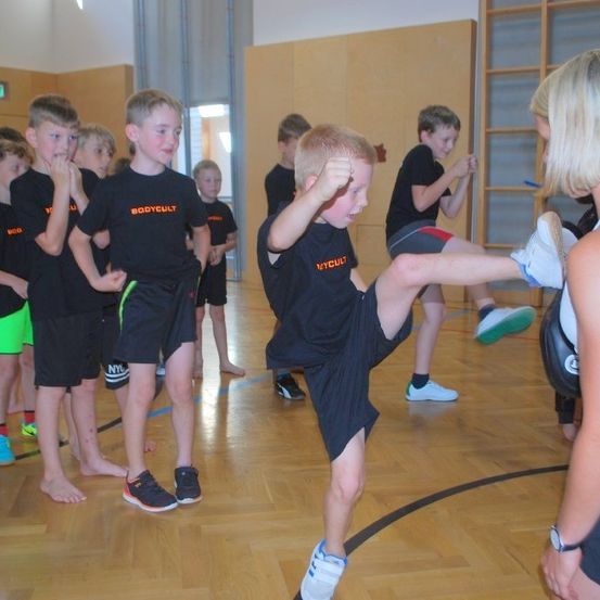 Eine Gruppe junger Jungen in einem Fitnessstudio lernt Kampfkünste von einer Frau. Sie sind in schwarzen und orangefarbenen T-Shirts mit dem Wort 'Bodycult' gekleidet.