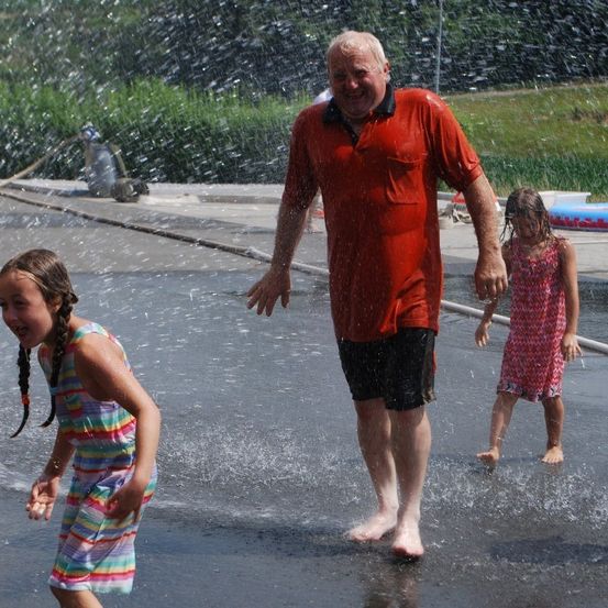 Ein älterer Mann in einem roten T-Shirt spielt mit zwei kleinen Mädchen in einem Wasserbrunnen.