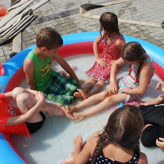Mehrere Kinder versammeln sich um einen kleinen aufblasbaren Pool, möglicherweise spielen sie darin. Ein Junge sitzt mit den Beinen im Pool.