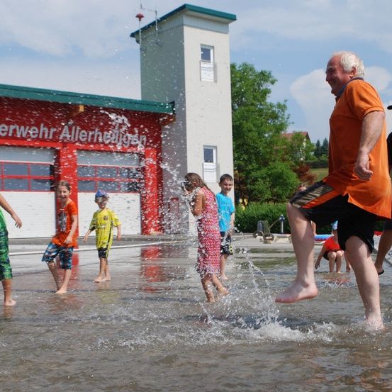 Mehrere Kinder und Erwachsene spielen in einem Brunnen vor einer Feuerwache. Das Gebäude hat rote Wände und ein grünes Dach.