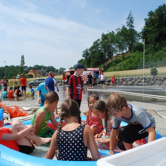 Kinder spielen in einem großen aufblasbaren Pool im Freien. Einige sitzen, andere stehen und genießen das Wasser. Ein Feuerwehrwagen ist in der Nähe geparkt.