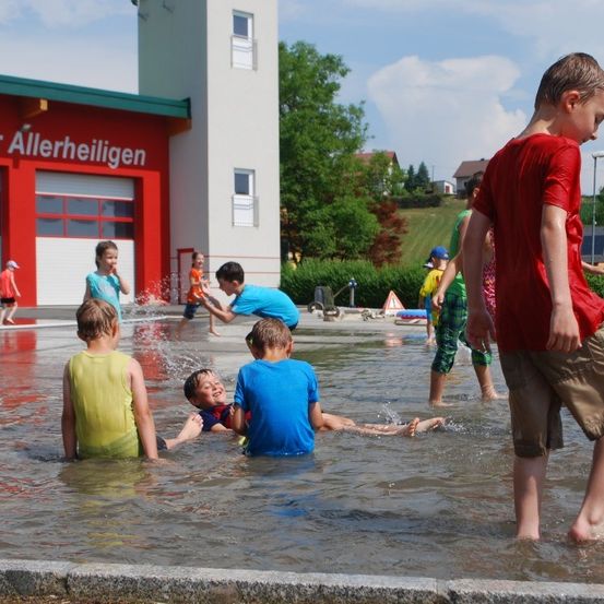 Kinder spielen in einer Wasserfläche vor einer Feuerwehrstation. Einige stehen im Wasser, während andere herumspritzen.