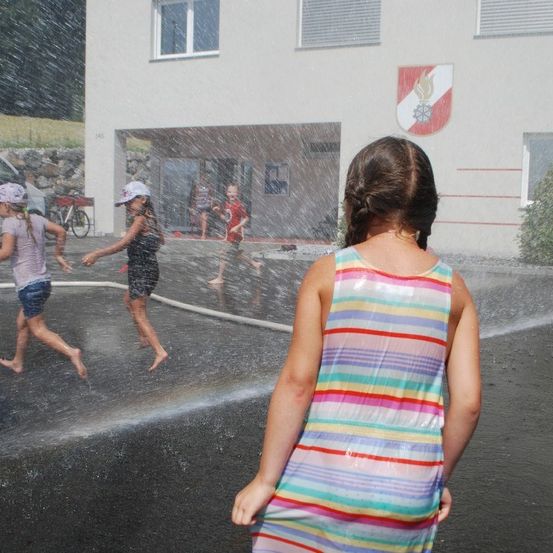 Kinder spielen in einem Wasserbrunnen vor einem Gebäude mit einem Feueremblem, ein Kind beobachtet von hinten.