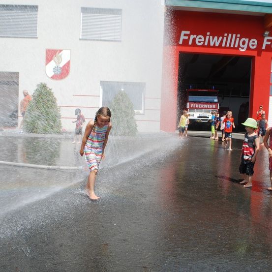 Kinder spielen und planschen in Wasserbrunnen vor einer Feuerwache. Die Wache hat ein rotes Äußeres und ein Logo an der Wand.