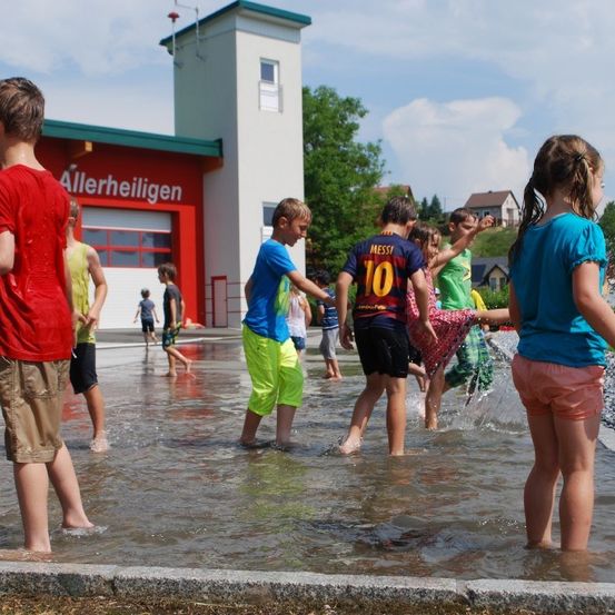 Mehrere Kinder spielen im Wasser vor einem Gebäude mit der Aufschrift Allerheinligen.